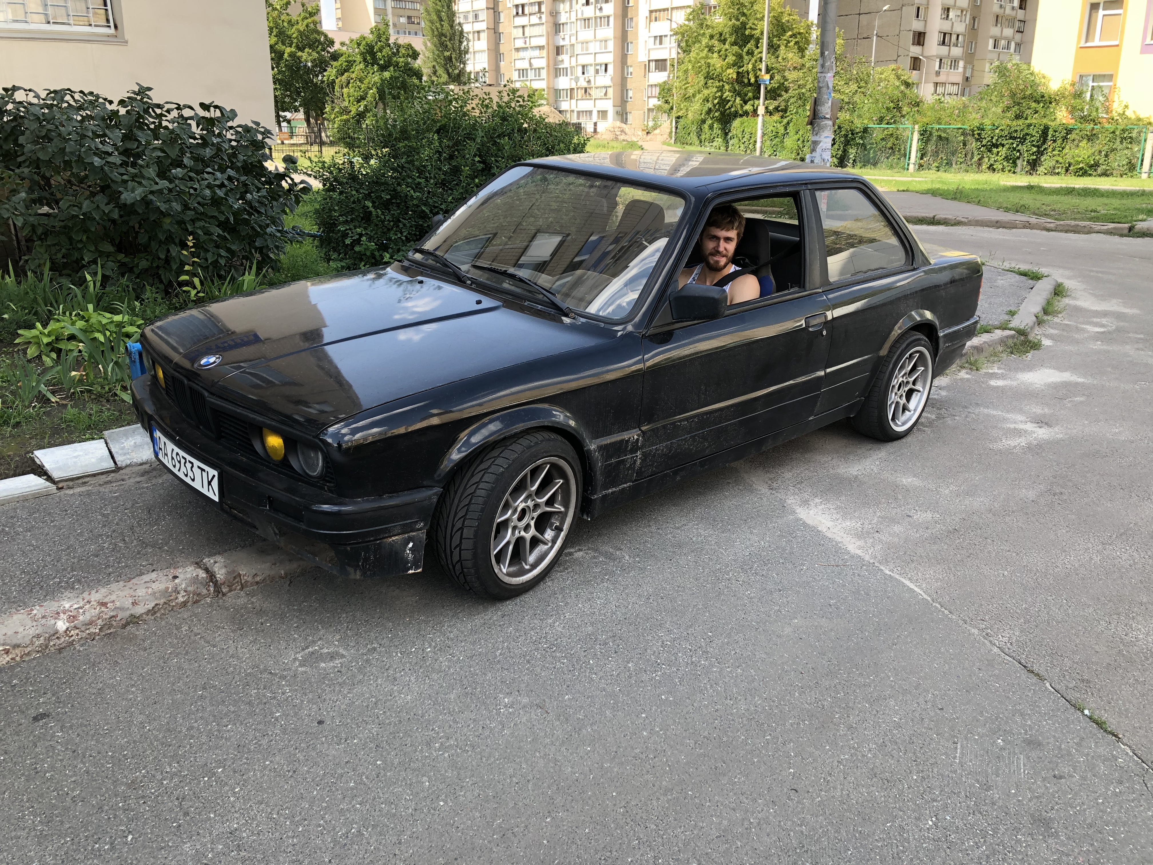 Yaroslav sitting in his black BMW E30 318 coupe on a summer street